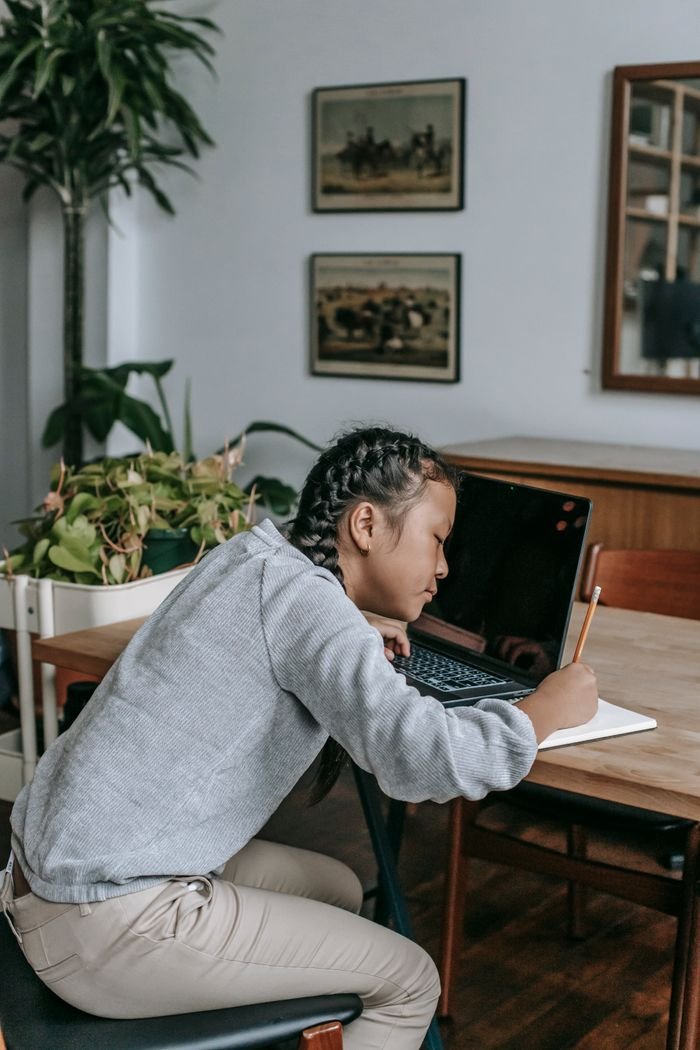 Side view of concentrated Asian girl in casual clothes sitting at table with opened laptop and writing in copybook