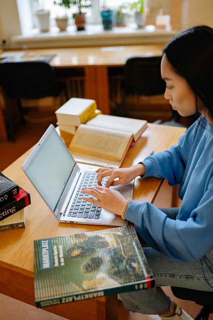 A young woman is focused on studying with her laptop at a wooden desk with open books.